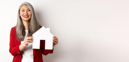 Asian female real estate agent showing paper house cutout, broker smiling friendly and selling property, standing over white background photo