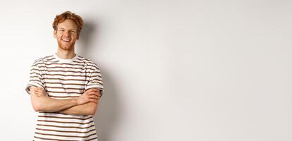 Happy redhead guy with bristle laughing and smiling at camera, standing with hands crossed on chest over white background photo