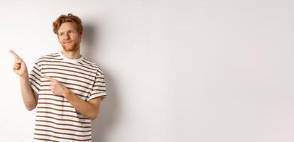 Doubtful young man with bristle and red hair looking, pointing fingers left at something average, staring skeptical at promo, white background photo