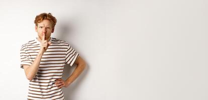 Angry redhead man looking displeased and hushing at camera, making shh gesture, standing over white background photo