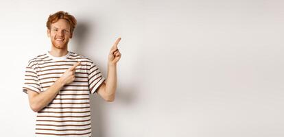 Handsome redhead man showing logo, pointing fingers right and smiling, standing over white background photo