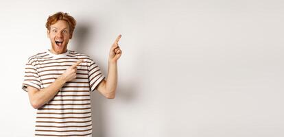 Excited redhead guy in striped t-shirt checking out special deal, pointing fingers right and smiling at camera, white background photo