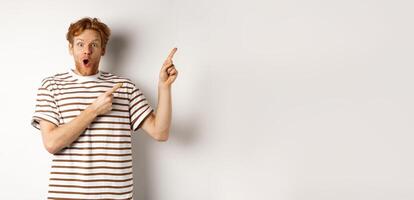Excited man with curly red hair showing promo offer and looking amazed, pointing fingers at upper right corner logo, white background photo