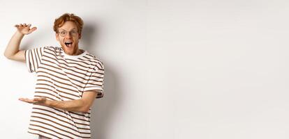 Amazed redhead man in glasses showing something large size and smiling at camera, saying wow, standing over white background photo
