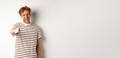Image of funny redhead guy in glasses laughing at you, pointing finger camera and making fun of someone, standing over white background photo
