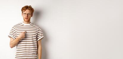 Confused redhead man in glasses pointing at himself, looking questioned at camera, white background photo