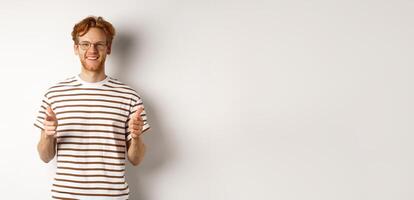 Young man with red hair and glasses pointing fingers at camera and smiling, need you, standing over white background photo