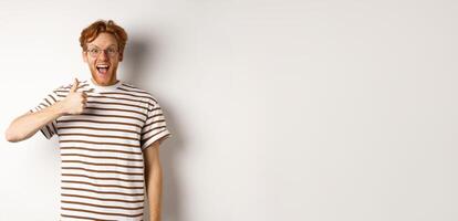Amazed and fascinated redhead guy checking out something awesome, showing thumb-up and scream yes with joy, standing over white background photo