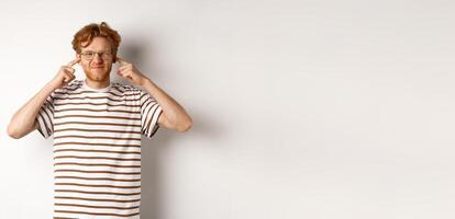 Annoyed young man complaining on loud noise, shut ears and grimacing from discomfort, standing over white background photo