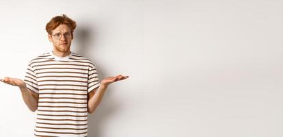 Confused boyfriend with red hair shrugging, spread hands sideways and staring at camera puzzled, asking what, stnading over white background photo