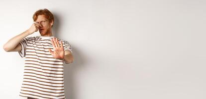 Young man with red hair staring at camera and shut nose from bad smell, looking disgusted and refusing, white background photo