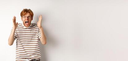 Angry young man with red hair shouting at camera, screaming and looking outraged, shaking hands, standing over white background photo