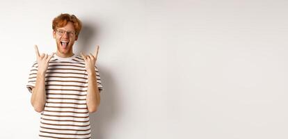 Funny and happy redhead man having fun, showing rock-n-roll horn and sticking tongue, enjoying party, standing over white background photo