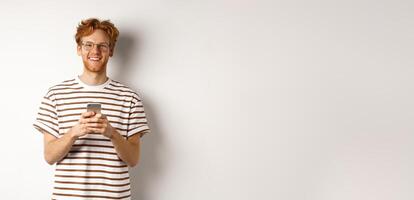 Technology and e-commerce concept. Redhead guy in glasses using mobile phone and smiling. Young man with smartphone staring happy at camera, white background photo