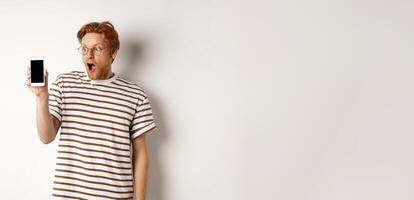 Technology and e-commerce concept. Surprised and shocked redhead guy checking out online promotion, showing blank smartphone screen and drop jaw, white background photo