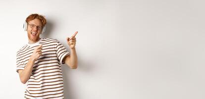 Technology concept. Happy redhead man listening music in headphones and dancing and pointing fingers right, standing over white background photo