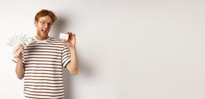 Shopping and finance concept. Young redhead man in glasses choosing plastic credit card, showing dollars and smiling, standing over white background photo