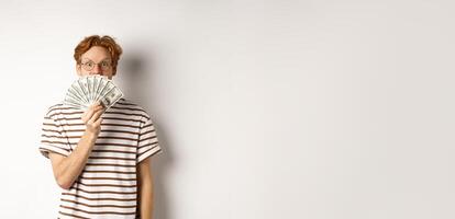 Shopping and finance concept. Lucky redhead guy winning, showing prize money and smiling happy at camera, standing in glasses and t-shirt over white background photo