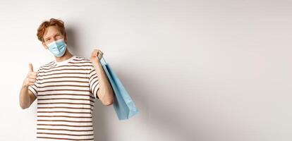 Concept of covid-19 and shopping. Satisfied young man looking pleased after shopping, wearing face mask, showing thumbs-up, recommend store, white background photo