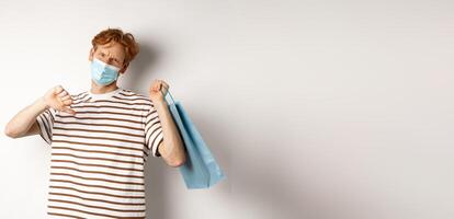 Concept of social distancing and shopping. Disappointed young man with red hair, wearing face mask, holding shopping bag and showing thumb-down from dislike, disapprove store photo