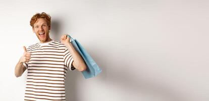 Happy young man with red hair, shopping in stores, showing thumbs-up and holding paper bag over shoulder, recommending shop, white background photo