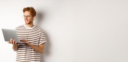 Young redhead freelancer working on laptop, typing on computer keyboard and smiling, standing over white background photo