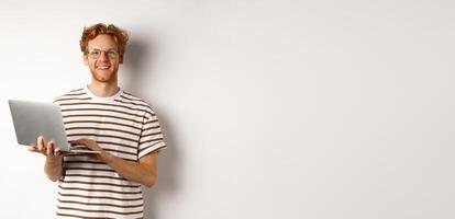 Smiling young man working on laptop and looking joyful, standing over white background photo