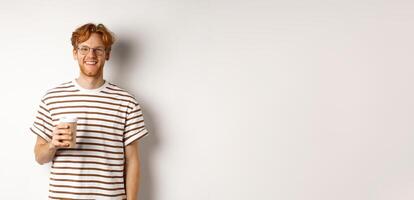 Handsome young man with beard and red messy hair, wearing glasses with striped t-shirt, drinking coffee from takeaway and smiling, white background photo