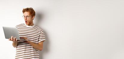 Wow, check this out. Excited redhead guy in glasses staring at laptop screen, reading online promo offer with amazed face, white background photo