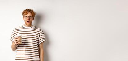 Amazed redhead man in glasses holding coffee cup and staring at camera with complete disbelief, standing in striped t-shirt against white background photo