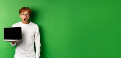 Impressed redhead guy in glasses drop jaw after seeing online promo, showing laptop screen and staring at camera with disbelief, standing over green background photo