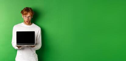 Impressed redhead nerdy guy in glasses showing blank laptop screen and looking at display amazed, standing over green background photo