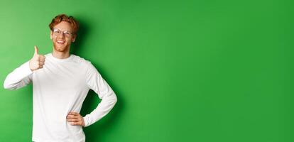 Cheerful young man with beard and red hair, wearing glasses, showing thumb-up in approval and smiling, say yes, standing over green background photo