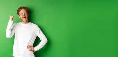 Young cheerful guy scream of joy, winning prize and celebrating, making fist pump gesture and smiling happy, standing over green background photo
