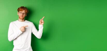 Skeptical caucasian man in glasses having doubts, pouting and frowning while pointing fingers right at something bad, standing over green background photo