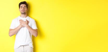 Romantic and nostalgic man holding hands on heart, close eyes and remember something, standing over yellow background photo