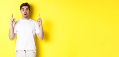 Surprised handsome guy in white t-shirt, pointing fingers up, interested about advertisement, standing against yellow background photo