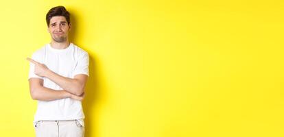 Skeptical and reluctant guy in white t-shirt, pointing finger left and smirking unamused, showing bad advertisement, standing over yellow background photo