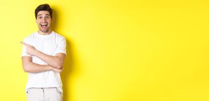 Happy man pointing finger left, showing advertisement on copy space, smiling amused, standing in white clothes against yellow background photo