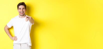 Confident smiling man pointing at you camera, standing in white clothes against yellow background photo