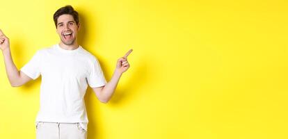 Happy stylish guy showing two variants, pointing fingers sideways at left and right promo, standing over yellow background photo