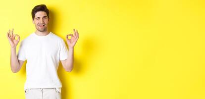Confident handsome man winking, showing okay signs in approval, like something good, standing over yellow background photo