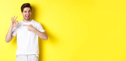 Image of handsome young man approve something, showing okay sign and winking, standing against yellow background photo