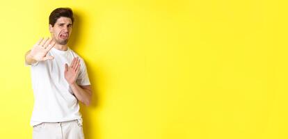 Disgusted man refusing, grimacing from dislike and aversion, begging to stop, standing in white t-shirt against yellow background photo