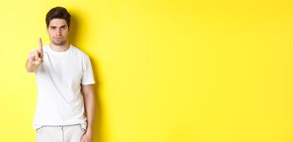 Confident man saying no, decline and prohibit something, showing one finger and frowning, standing over yellow background photo