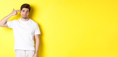 Bothered man shooting himself with finger gun, looking displeased and tired, standing against yellow background photo