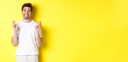 Hopeful smiling man holding fingers crossed, making wish or praying, standing over yellow background photo
