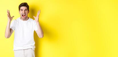 Angry man yelling and shaking hands, grimacing with hatred, standing hateful against yellow background photo