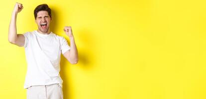 Excited handsome man winning and shouting for joy, raising hand up and triumping, achieve goal, standing over yellow background photo