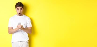 Man looking surprised in smartphone, reading message on cell phone, standing in white outfit against yellow background photo